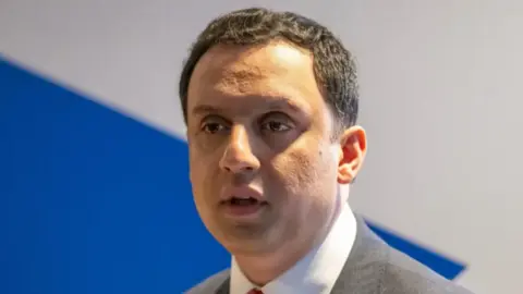 PA Media Anas Sarwar is wearing a grey suit and red tie and is standing in front of a blue and white background. 
