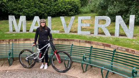 Harriet Harnden wearing a black top and bottoms holding her mountain bike stood in front of a Malvern sign