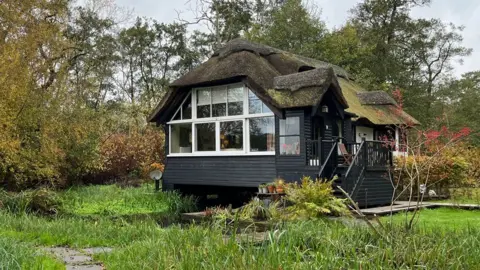 Shaun Whitmore/BBC A black timber building with a mossy thatched roof and white windows. The building is built over a river. It is surrounded by grass with trees in the background 