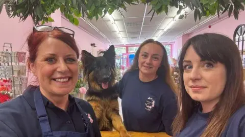 Enchanted Rose Three florists and a dog take a selfie at the counter of a flower shop