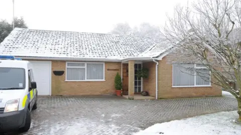 The Cambrisshire police stood on one side with a police vehicle with a bungalow. It is winter and there is a ice cover in the garden of the bungalow.