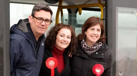 Andy Burnham at a Labour campaign event with its candidate Angeliki Stogia and Deputy Leader Lucy Powell - all three are standing in the entrance of a campaign bus, with the two women wearing red rosettes