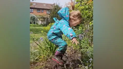 Wicksteed Park A girl in a blue coat and black wellington boots leaps into a muddy puddle. The droplets from the splash fill the surrounding area