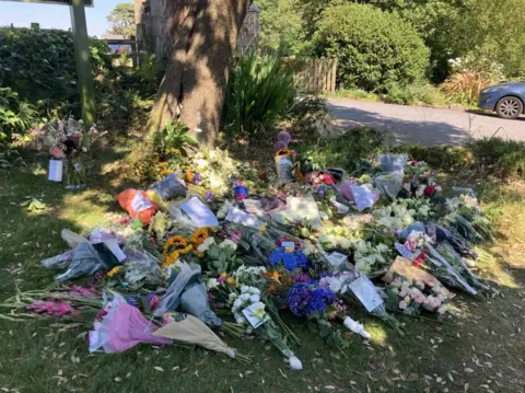 Dozens of bouquets of flowers laid on the grass in front of a tree near a hotel. Among the floral tributes at the back is a black and white photo of Widdecombe.