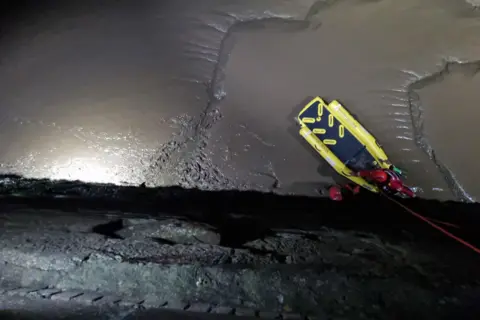 Maryport Inshore Rescue Two rescuers on fluorescent yellow inflatable lifeboat are pulled up at a bottom of a grey stone wall. The scene is lit by torch. 