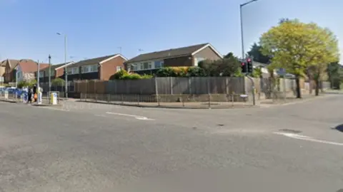 The corner of a road junction. There are road marking showing turning lanes on the ground and a built up area of houses. 