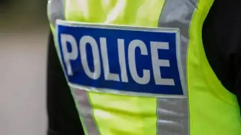 A generic police jacket. The officer is wearing a black jacket and a hi-vis overall which is yellow or green with grey stripes. The word POLICE is printed in capital letters in white on a blue background on the back of the jacket
