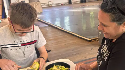 Nancy working with one of her students - she is wearing sunglasses on the top of her head and a black t-shirt. The pupil is scooping avocado and wearing a grey T-shirt.