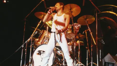 Getty Images Freddie Mercury standing in front of a drumkit as he sings into a microphone on stage during a live concert performance at the National Bowl in Milton Keynes