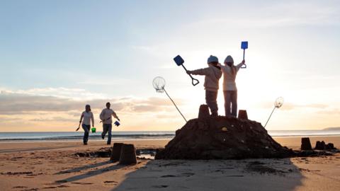 Two small children stand on a large sandcastle on a beach at sundet, holding their spades aloft. Their parents run towards them from the direction of the sea.