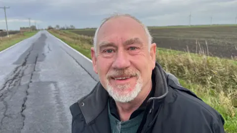 John Devine/BBC Mike Swann, 63, has short grey hair on the very top and sides of his head, and a neat grey beard and moustache, he is wearing a black padded jacket and a green round collared jumper underneath with a black shirt under that.
Behind him is a very badly cracked rural road, with farmers fields either side, no white lines, no lighting.