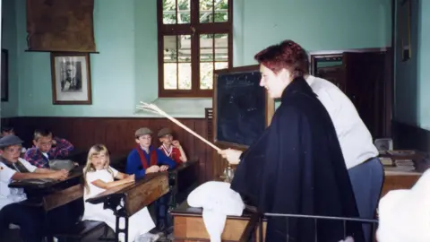 Anton Honchanka A woman stands at the front of an old classroom with a chalkboard behind her. Children, including Anton, sit at tables watching her.