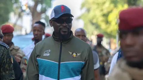 Guinea Presidency Mamadi Doumbouya seen outside wearing a tracksuit, baseball cap and dark glasses. A soldier in a red beret is seen on the right in the foreground out of focus.