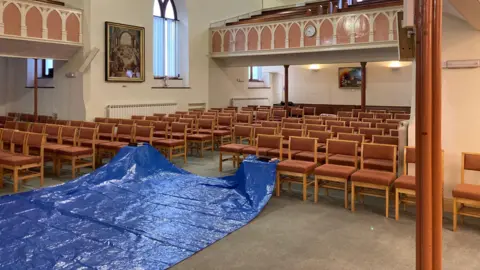 The Main Church hall. There is a large blue tarp on the floor and in the background there are rows or chairs - two of which are covered by the tarp 