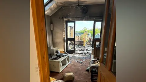 SFRS A living room that is black with fire and there are broken and burnt items scattered across the floor. Through some double doors is an area where a conservatory should be. There are no windows and there is glass and blackened debris on the floor, with a firefighter looking at it
