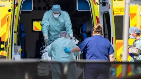 Getty Images Two medical workers in full PPE - green overwear, hat and masks, helping someone in a wheelchair into the back of an ambulance. A nurse in normal scrubs and mask is helping them.