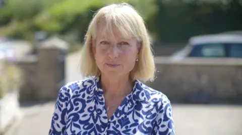 Jamie Niblock/BBC A woman stands outside on a sunny day. She has blonde hair that has been cut in a bob style. She wears a white blouse with blue floral patterns on it. 