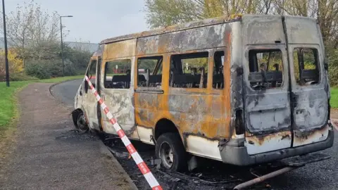 A burnt-out bus on a road. Tape is around the bus. which is scorched and rust-coloured, with just some white paint remaining. There is debris on the road and, just visible on the bus, there is there is faint writing of the name of the school: Caroline Haslett Primary. 