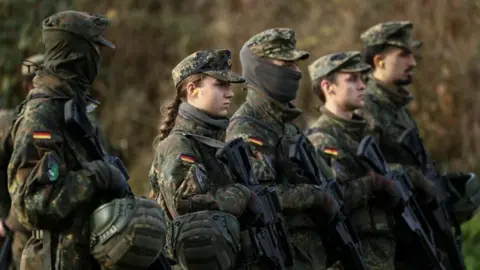 Reuters Soldiers and soldiers stand with rifles in a field, wearing camouflage clothing with the German flag sewn on their upper arms.