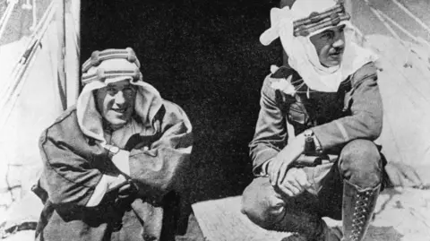 Bettmann via Getty Images A black and white image of two men kneeling down. They are wearing military uniform underneath typical Arab clothing.