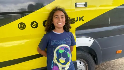 A young boy with long brown curly hair, wearing a football t-shirt is leaning on a bright yellow van, smiling at the camera.