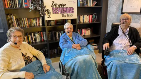 Left Maureen , Ann in the middle and Doreen on the right. Three elderly women sat in wheelchairs with blue blankets over them. They have hands free microphones on. Behind them there is a book shelf with a sign that reads 'Do you remember the time?'