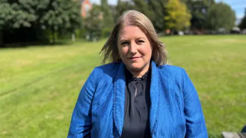 A lady with blonge hair, wearing a navy dress and a royal blue blazer, standing on a grassy area with trees in soft focus behind her.