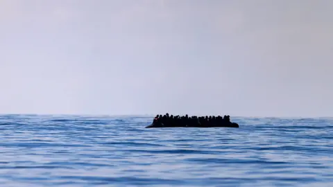 Getty Images A inflatable dinghy carrying around 65 migrants can be seen silhouetted against a grey sky on a blue sea, as it crosses the English Channel in March