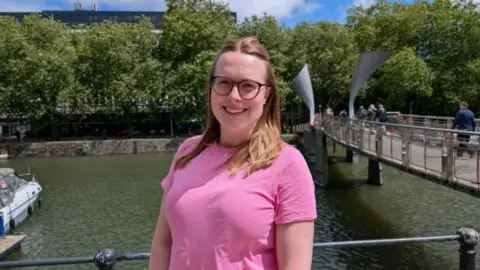 Handout Lisa Armstrong, a woman with glasses and long brown hair is wearing a pink top as she poses next to Pero's Bridge in Bristol Harbour
