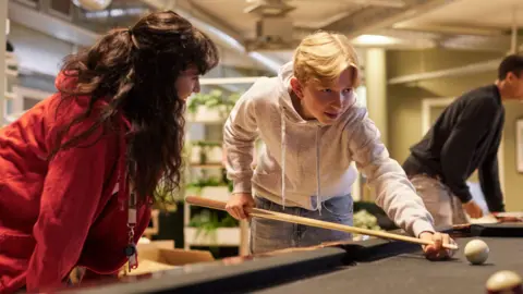 Getty Images A boy playing pool. He wears a grey hoody and jeans. A girl stands to his left in a red hoody.