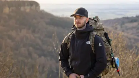 A man stands in rural terrain. There are hills in the background. He wears a black jacket and baseball cap, and carries a large, military-style backpack.