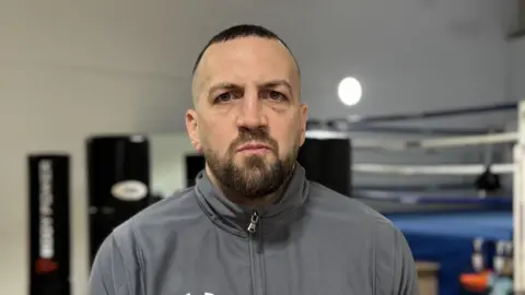 Man in his 30's wearing a grey Hurley zip up stood in a gym. He has short hair and a full beard. Behind him you can see a boxing ring.