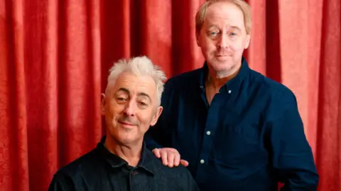 Tommy Ga Ken Wan Two men photographed against a red curtain. One has receding red hair and is wearing a navy shirt. He has his hand on the other's man's shoulder. The second man is wearing a dark tartan shirt and has grey hair styled in a quiff. Both men have a slight grin on their faces.