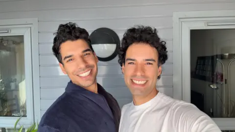 Twin 30-something brothers Riccardo and Simone, with dark brown hair and dark brown eyes, stand next to each other and grin at the camera 