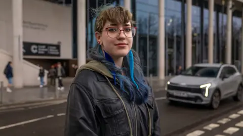 A girl with blue hair and blue feather earrings smiling faintly at the camera. She wears red glasses and a black leather jacket. She's standing in front of a modern glass building in front of a road.