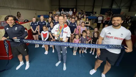 Two men hold a blue ribbon that says Stroke Trampoline Gymnastics Club Grand Opening, in the middle Bryony Page, who is wearing a white top and her Olympic medical, stands smiling while holding scissors. Behind are her dozens of children and adults of mixed ages
