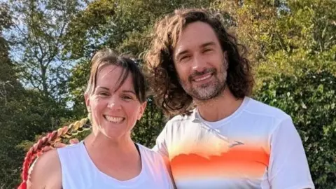 Carly Vine A woman - Carly Vine - standing next to celebrity fitness coach Joe Wicks. Both are smiling directly at the camera, standing in a park and wearing running gear.