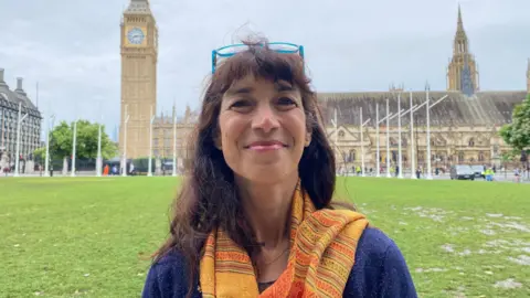 A woman stands in front of the Houses of Parliament. She has brown hair, a blue jumper and an orange scarf. She has glasses perched on her head.