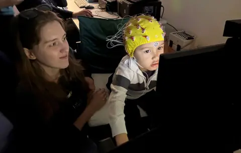A young boy wearing a yellow cap fitted with multiple wires sits at a computer workstation and watches a video while engaged in a research activity. Behind, computer equipment and monitors fill the dimly lit room and one of the researchers looks on in frame.