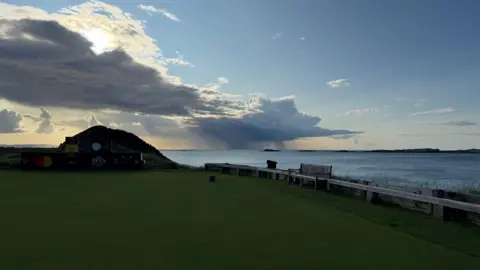 Dark grey clouds covering a golf course leaving a dark shadow. The sea is right next to the course.