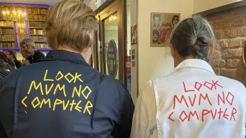 A man and a woman with their backs to the camera. He is to the left and has brown hair, she has grey hair tied back in a plait. They are both wearing boiler suits - his is navy blue with yellow lettering across the back, hers is white with red lettering. The lettering reads LOOK MUM NO COMPUTER 