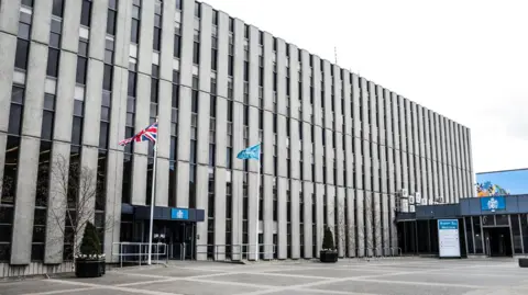 The outside of Darlington Town Hall which is a large, brutalist building with columns of narrow windows. There is a Union Jack flag flying outside the entrance along with another blue flag. 