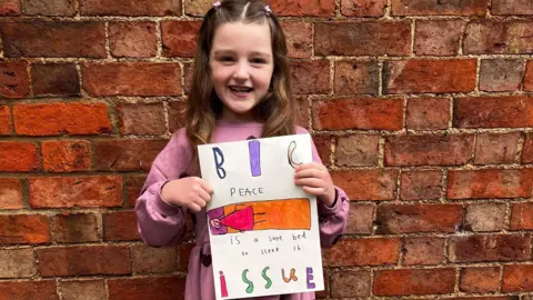 April is standing in front of a brick wall smiling at the camera. She has long brown hair and is wearing a pink dress. She is holding a copy of her drawing which says Big Issue on it and shows a girl sleeping in a bed.