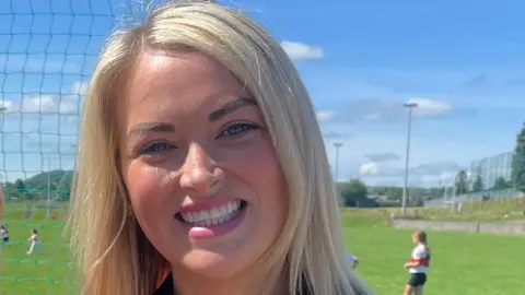 Ciara is smiling at the camera. There are children playing GAA behind her