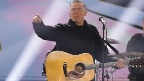Bryan Adams on stage with a drum kit and blue and purple backdrop behind him, and a microphone in front of him. He is pointing towards the audience, has oiled-back hair and is wearing a black shirt.  