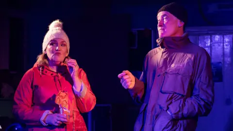 Origin8Photography Two stage actors performing a play. They are dressed in coats and beanie hats. A woman is eating biscuits while a man next to her is talking.