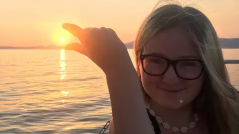 Lucy is wearing glasses and a necklace, posing as if she is trying to pinch the sun at sunset