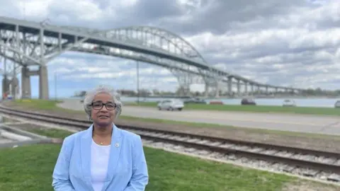 Woman in light blue blazer stands in front of bridge