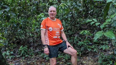 Paul Gentry Paul Gentry indossa una maglietta arancione di beneficenza e pantaloncini scuri e guarda nella telecamera mentre si trova nel bosco