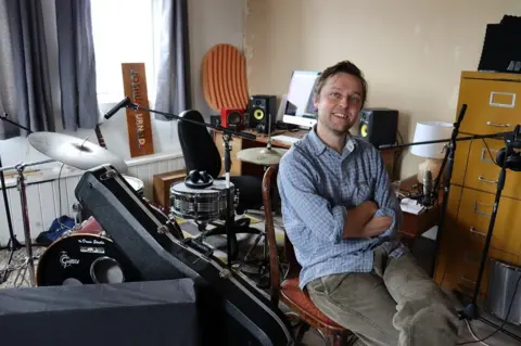 Joshua Burnside in grey trousers and a blue check shirt, pictured in front of drums, a computer and other musical equipment